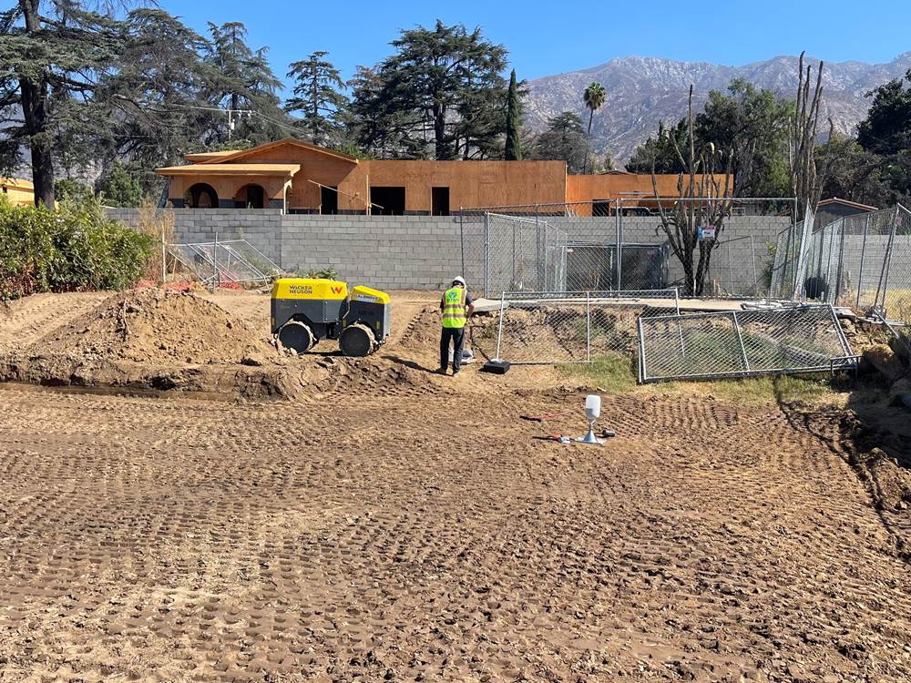 Titan technician in a safety vest on a graded residential building pad beside a trench roller, with a sand-cone density test set up in the foreground