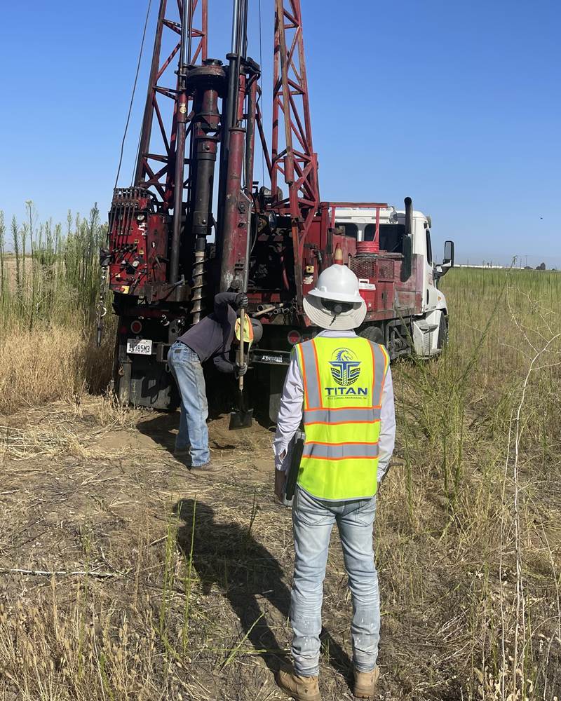 Titan Geotechnical field engineer in a branded safety vest and hard hat overseeing a truck-mounted drill rig as the crew advances a boring in an open field
