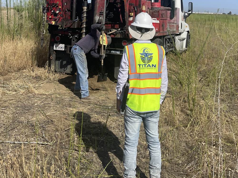 Titan Geotechnical field engineer in a branded safety vest and hard hat overseeing a truck-mounted drill rig as the crew advances a boring in an open field
